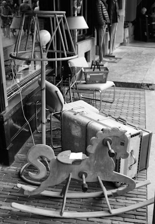 A collection of vintage items arranged outdoors along a sidewalk, including a large, old-fashioned suitcase, a wooden rocking horse, and a variety of lamps. Behind these items, a shop window and entrance are visible, with reflections of passersby on the glass. The scene has a nostalgic, antique feel.