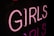 Neon pink letters spell out the word 'GIRLS' against a dark background, casting a soft glow around them.