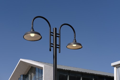 A modern street lamp with two bulbs mounted on a curved, minimalist pole set against a clear blue sky. Below the lamp, a part of a modern building with a slanted roof and large glass windows is visible.