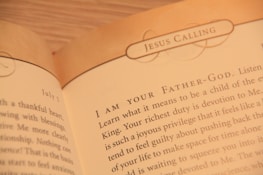 A close-up of an open book displaying text with the heading 'JESUS CALLING'. The text appears to be a religious or spiritual message, with focus on phrases like 'I AM YOUR FATHER-GOD' and discussion of devotion and spiritual duties.