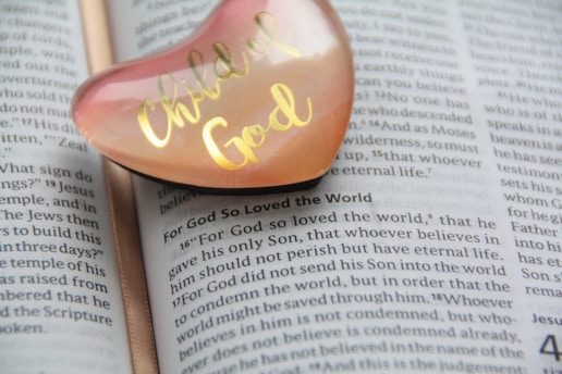 A heart-shaped object with 'Child of God' written in gold is placed on an open page of a Bible. The focus is on the text that includes the passage 'For God so loved the world,' with the heart object slightly transparent, showing a peach and pink hue.