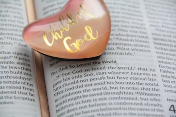 A heart-shaped object with 'Child of God' written in gold is placed on an open page of a Bible. The focus is on the text that includes the passage 'For God so loved the world,' with the heart object slightly transparent, showing a peach and pink hue.