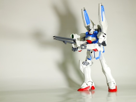 A highly detailed model of a robot stands prominently with a futuristic design. It features a combination of sleek, angular components, primarily in white with accents of blue, red, and gray. The robot is posed in an action stance, holding a large weapon, highlighting its mechanical and battle-ready features. The background is plain and unobtrusive, drawing focus to the intricacies of the robot.
