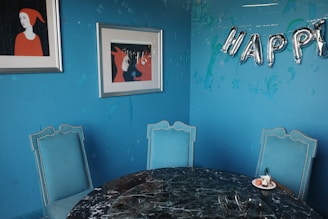 A stylish interior with a blue-painted wall adorned with two framed artworks featuring figures in red clothing. The wall hosts metallic balloon letters spelling 'HAPPY'. Below, a round, dark marble table is surrounded by three blue upholstered chairs, with one holding a small plate with dessert.