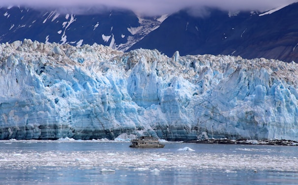 A vast and towering glacier dominates the scene, with jagged and rugged ice formations in shades of blue and white. In the foreground, a small boat traverses the icy waters, providing a sense of scale against the immense glacier. The backdrop features dark, snow-capped mountains partially obscured by clouds.