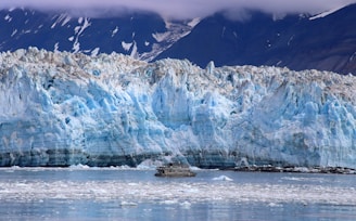 A vast and towering glacier dominates the scene, with jagged and rugged ice formations in shades of blue and white. In the foreground, a small boat traverses the icy waters, providing a sense of scale against the immense glacier. The backdrop features dark, snow-capped mountains partially obscured by clouds.