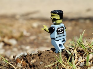 A toy figure resembling a rock and roll-themed monster is positioned on a background of dirt and grass. It has a light green face with black hair and is wearing a shirt labeled 'Shock 'n' Roll.' The scene is outdoors with a blurred background, giving focus to the toy.