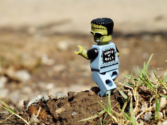 A toy figure resembling a rock and roll-themed monster is positioned on a background of dirt and grass. It has a light green face with black hair and is wearing a shirt labeled 'Shock 'n' Roll.' The scene is outdoors with a blurred background, giving focus to the toy.