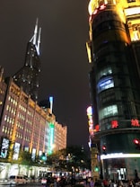 A nighttime cityscape with tall buildings illuminated by bright lights. There's a distinctive skyscraper with a spire, and several signs with colorful neon lights in Chinese characters. The street below is busy with traffic and pedestrians, while trees line the sidewalks.
