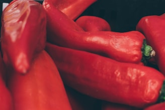 A close-up view of a pile of long, red peppers with smooth, shiny skins. The peppers are vibrant in color, showing varying shades of red, with small green stems visible on some.