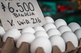 A carton of white eggs is prominently displayed with a handwritten sign indicating the price and quantity. The background features a blurred market setting with various produce.