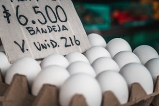 A carton of white eggs is prominently displayed with a handwritten sign indicating the price and quantity. The background features a blurred market setting with various produce.