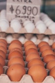 Brown eggs neatly arranged in a carton tray with a second tray of white eggs in the background. A blurred price sign is visible above the eggs.