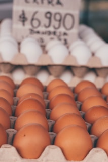 Brown eggs neatly arranged in a carton tray with a second tray of white eggs in the background. A blurred price sign is visible above the eggs.
