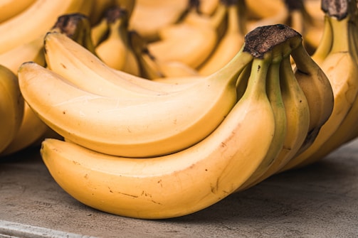 A close-up view of a bunch of ripe yellow bananas with a few blemishes, resting on a stone or concrete surface. The bananas have a thick peel and some dark brown tips.