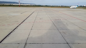 An airplane is parked on an expansive concrete airport tarmac. Red and black lines mark different areas of the tarmac. In the background, there is a city skyline with buildings and a green area separating the airport from the urban area.