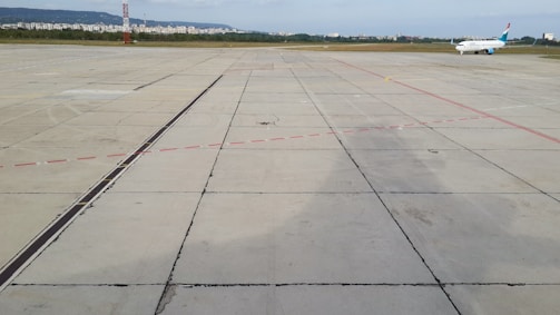 An airplane is parked on an expansive concrete airport tarmac. Red and black lines mark different areas of the tarmac. In the background, there is a city skyline with buildings and a green area separating the airport from the urban area.