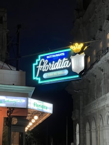Neon signage for a bar and restaurant named Floridita is prominently displayed against a nighttime backdrop. The sign is illuminated in bright colors, with the words 'Restaurante Bar Floridita' and a line underneath reading 'La Cuna del Daiquiri.' An additional smaller illuminated sign with a white background can be seen below, referencing Ernest Hemingway. Architectural details of a building with arched windows are visible on the right.