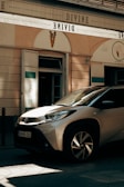A beige compact car is parked on a shadowed street in front of a small café or boutique. The signage above, which includes the word 'DIVINE,' is written in a stylish font. The exterior walls of the building are cream-colored with subtle architectural details, and a bundle of grapes decorates the upper center facade.