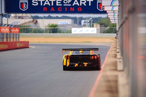 A racing car speeds along a race track, emitting flames from its exhaust as it drives past barriers advertising 'uhoda'. Above the track, a banner reads 'STATE OF ART RACING'. The track curves gently and is surrounded by barriers and buildings in the distance.