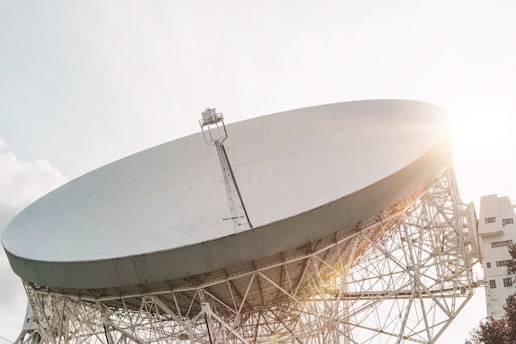 A large radio telescope dish with a sturdy metal framework, positioned at an angle under bright sunlight. The structure is massive and the sun is shining brightly on its surface, creating lens flare effects. The sky is clear with minimal cloud presence.