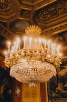 A luxurious, ornate chandelier with intricate gold detailing and crystal accents hangs from an elaborately decorated ceiling. The chandelier is lit with numerous bulbs, casting a warm glow that reflects off the gold and crystals. The ceiling features ornate moldings and patterns that add to the opulence of the setting. Rich textures and dark tones are present in the surrounding decor, suggesting a lavish interior space.