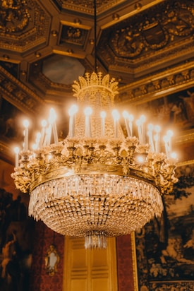 A luxurious, ornate chandelier with intricate gold detailing and crystal accents hangs from an elaborately decorated ceiling. The chandelier is lit with numerous bulbs, casting a warm glow that reflects off the gold and crystals. The ceiling features ornate moldings and patterns that add to the opulence of the setting. Rich textures and dark tones are present in the surrounding decor, suggesting a lavish interior space.