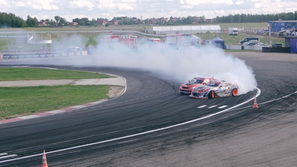 Two sports cars are drifting side by side around a curved track, producing large plumes of smoke from their tires. The track is marked with tire marks from previous drifts and is bordered by grass. The background includes some structures, scattered cars, and trees under a partly cloudy sky.