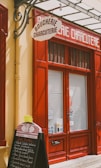 A storefront with vibrant red doors and window frames, featuring a traditional butcher shop sign that reads 'Boucherie Charcuterie' in French. A chalkboard menu is displayed outside, listing various offerings. The atmosphere suggests a quaint, classic European shop.