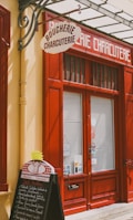 A storefront with vibrant red doors and window frames, featuring a traditional butcher shop sign that reads 'Boucherie Charcuterie' in French. A chalkboard menu is displayed outside, listing various offerings. The atmosphere suggests a quaint, classic European shop.
