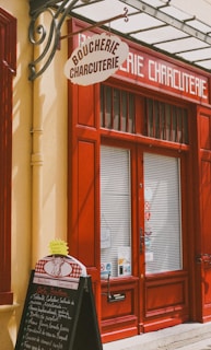 A storefront with vibrant red doors and window frames, featuring a traditional butcher shop sign that reads 'Boucherie Charcuterie' in French. A chalkboard menu is displayed outside, listing various offerings. The atmosphere suggests a quaint, classic European shop.