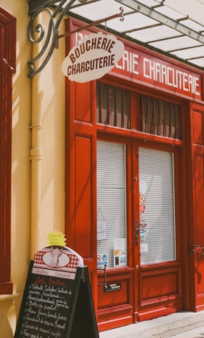 A storefront with vibrant red doors and window frames, featuring a traditional butcher shop sign that reads 'Boucherie Charcuterie' in French. A chalkboard menu is displayed outside, listing various offerings. The atmosphere suggests a quaint, classic European shop.