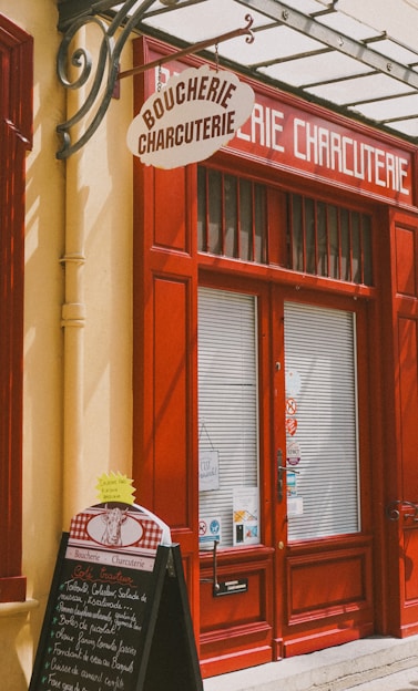 A storefront with vibrant red doors and window frames, featuring a traditional butcher shop sign that reads 'Boucherie Charcuterie' in French. A chalkboard menu is displayed outside, listing various offerings. The atmosphere suggests a quaint, classic European shop.