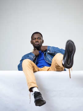 A person is sitting casually with one leg raised, wearing a blue shirt, denim jacket, tan pants, and black shoes. The background is plain and white, adding a stark contrast to the outfit.