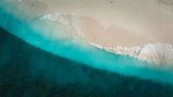 Aerial view of turquoise ocean meeting white sandy beach under bright sunlight.