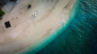 An aerial view of a sandy beach with turquoise waves gently lapping the shore. Sparse human activity is present, with a few white chairs and a table set up near the water's edge. A small wooden structure or hut surrounded by greenery is visible on the left. The beach is mostly empty, suggesting a tranquil and serene atmosphere.