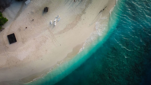 An aerial view of a sandy beach with turquoise waves gently lapping the shore. Sparse human activity is present, with a few white chairs and a table set up near the water's edge. A small wooden structure or hut surrounded by greenery is visible on the left. The beach is mostly empty, suggesting a tranquil and serene atmosphere.