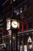 An illuminated steam-powered clock stands in front of a brick building at night. The clock is surrounded by clouds of steam, and its face is brightly lit, casting a warm glow. The building behind the clock is adorned with rows of windows and red accent lighting.