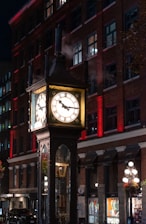 An illuminated steam-powered clock stands in front of a brick building at night. The clock is surrounded by clouds of steam, and its face is brightly lit, casting a warm glow. The building behind the clock is adorned with rows of windows and red accent lighting.