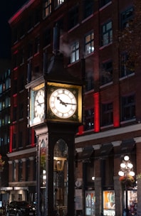 An illuminated steam-powered clock stands in front of a brick building at night. The clock is surrounded by clouds of steam, and its face is brightly lit, casting a warm glow. The building behind the clock is adorned with rows of windows and red accent lighting.