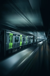 A sleek, modern train moves rapidly through a dimly lit underground station, creating a sense of speed and motion. The train features striking green and grey colors, with its headlights illuminating the path ahead.