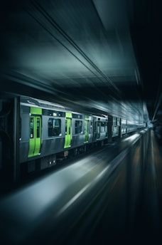 A sleek, modern train moves rapidly through a dimly lit underground station, creating a sense of speed and motion. The train features striking green and grey colors, with its headlights illuminating the path ahead.