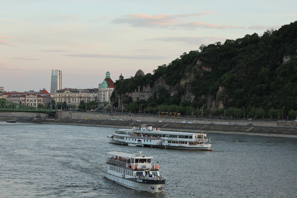 Two large white boats sail on a serene river with passengers enjoying their journey