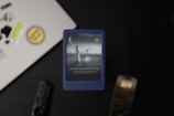 A Kindle device, displaying a grayscale image of a woman looking through a telescope at a large crescent moon, is placed on a surface. In the background, a laptop with various stickers, a remote control, and a smartphone are visible. The Kindle screen text reads 'Your next great read is waiting.'