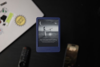 A Kindle device, displaying a grayscale image of a woman looking through a telescope at a large crescent moon, is placed on a surface. In the background, a laptop with various stickers, a remote control, and a smartphone are visible. The Kindle screen text reads 'Your next great read is waiting.'