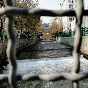 A river flows through a cityscape, framed by a metal fence. The water cascades over a small waterfall and continues between green-fenced stone paths lined with trees. In the background, urban buildings rise, partially obscured by leafless branches.