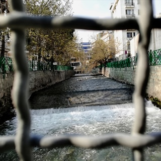 A river flows through a cityscape, framed by a metal fence. The water cascades over a small waterfall and continues between green-fenced stone paths lined with trees. In the background, urban buildings rise, partially obscured by leafless branches.