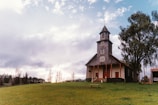 A rural, wooden church stands on a grassy hillside under a cloudy sky. The church features a tall steeple with a cross at the top and an inviting front porch with steps. A large tree grows close by, and a simple wooden bench sits in front of the building.