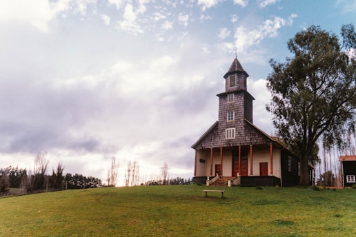 A rural, wooden church stands on a grassy hillside under a cloudy sky. The church features a tall steeple with a cross at the top and an inviting front porch with steps. A large tree grows close by, and a simple wooden bench sits in front of the building.