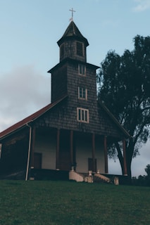 A rustic wooden church with a shingled roof stands on a grassy hill. It has a tall bell tower topped with a cross, and a large tree is visible to the side. The sky is cloudy, adding a serene and peaceful atmosphere.